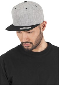 Cappello snapback di tessuto grigio con visiera nera, dotato di occhielli neri e design piatto. Indossato da una persona con una maglietta nera.