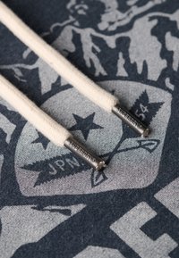 Lacci in cotone beige con estremità in metallo che presentano il marchio "SUPERDRY", abbinati a uno sfondo grafico scuro con motivi stellati.