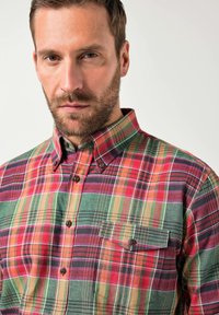 Uomo con capelli castani corti e barba che indossa una camicia a quadri multicolore rossa, verde e viola, abbottonata, con una tasca frontale.