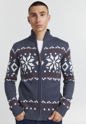 Dark blue zip-up sweater with a high collar, featuring white and brown snowflake patterns across the chest and sleeves. Ribbed cuffs and hem.