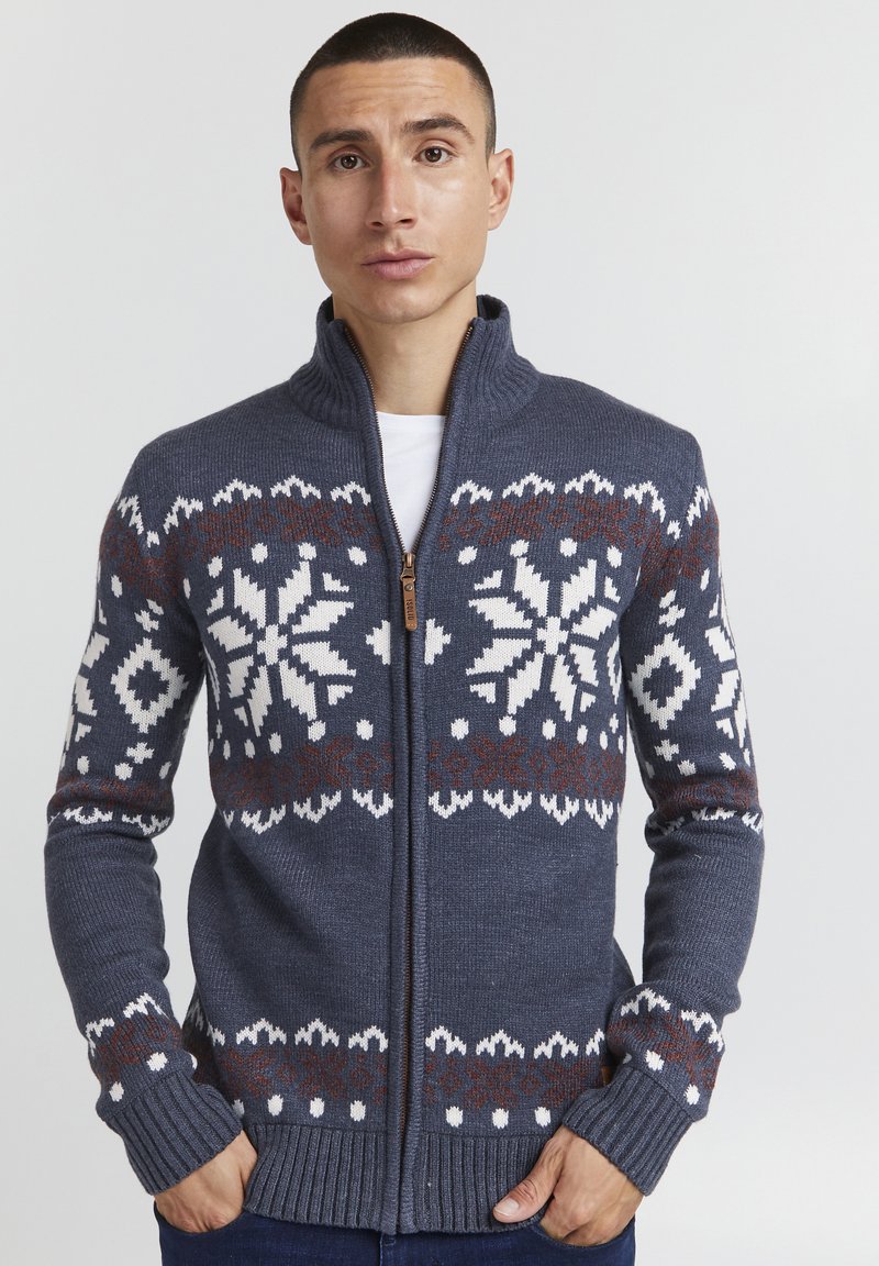 Dunkelblaues Zip-Up Sweatshirt mit hohem Kragen, das weiße und braune Schneeflockenmuster über der Brust und den Ärmeln aufweist. Gerippte Bündchen und Saum.