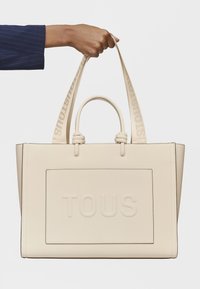 Sac fourre-tout beige avec logo "TOUS" en relief, texture lisse, deux anses supérieures et un design structuré. Les sangles présentent des détails de marque.