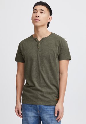 Oliven grønn kortermet t-skjorte med rund hals og tre knappestolper, laget av mykt, teksturert stoff. Brukt med blå jeans.
