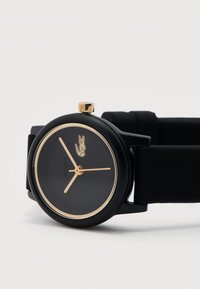 Μαύρο ρολόι χειρός Lacoste με χρυσές λεπτομέρειες, απλό καντράν, χρυστά δείκτες ώρας, λεπτών και δευτερολέπτων, και μαύρο λουράκι από υφασμένο σιλικόνη.