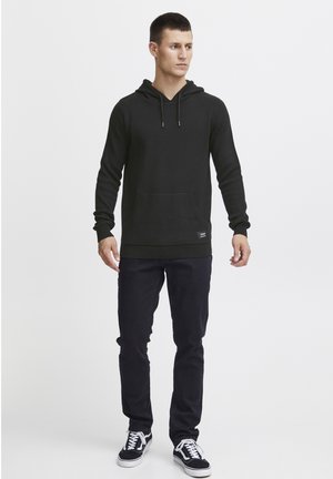 Black hoodie with a front pocket, drawstring hood, ribbed cuffs, paired with dark jeans and black sneakers. Simple, casual design.
