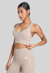 Beżowy stanik sportowy z dekoltem w kształcie łódki i plecami w stylu kolarskim, w zestawie z dopasowanymi leginsami o wysokim stanie. Posiada logo w kolorze białym.