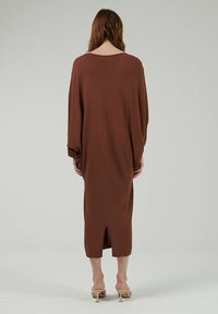 Vestido marrón de manga larga con corte holgado, de longitud a media pantorrilla y una abertura en la parte posterior para facilitar el movimiento. Textura de tela suave.