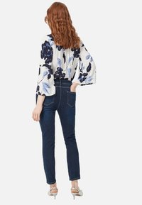 Mujer con cabello ondulado y rojo, vestida con una blusa blanca con estampado floral azul, jeans ajustados oscuros y sandalias de tacón blancas, de pie con la mano en la cadera.