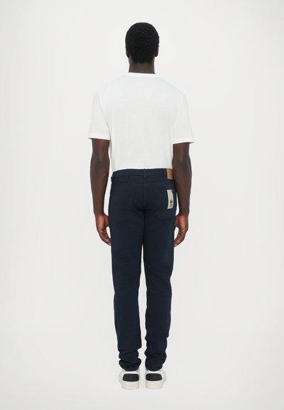 Jeans azul marino de corte ajustado, hechos de denim. Presentan un parche de cuero marrón en la parte trasera de la cintura y una etiqueta en el bolsillo trasero.