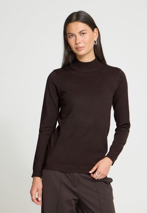 Femme aux longs cheveux foncés portant un pull à col roulé marron foncé et un pantalon assorti, posant avec une main dans la poche devant un fond uni.