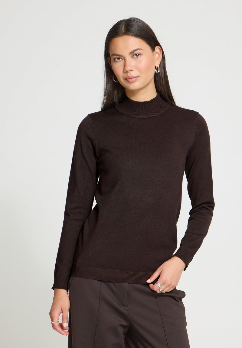 Femme aux longs cheveux foncés portant un pull à col roulé marron foncé et un pantalon assorti, posant avec une main dans la poche devant un fond uni.