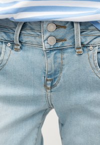 Τζιν παντελόνι από ανοιχτό μπλε τζιν με ψηλή μέση, διπλό κούμπωμα με μεταλλικά κουμπιά και ορατή ραφή, φορεμένο με μπλε και λευκή ριγέ μπλούζα.