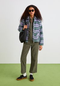 Fuzzy, purple and green plaid jacket with a black zipper; olive green jumpsuit; black bag; black sneakers with gum soles; round sunglasses.