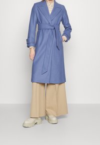 Femme portant un manteau lavande ceinturé, un pantalon beige à jambes larges, des chaussures chunky beiges et une chemise rayée glissée dans le manteau.