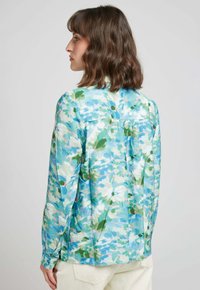 Blouse florale dans des tons de bleu et de vert avec un tissu texturé. Manches longues, empiècement au dos et ourlets fendus accentuent la coupe décontractée.
