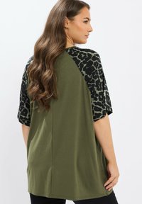 Vrouw met lang golvend bruin haar, gekleed in een olijfgroen top met zwarte en olijfgroene luipaardprintmouwen, staand tegen een effen witte achtergrond.