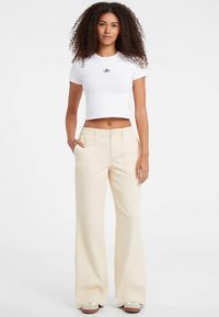White cropped t-shirt with a star emblem, paired with wide-leg, light beige pants. Model standing, hands in pockets, simplistic design.