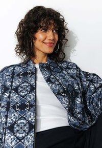 Veste bomber à motif bleu avec fermeture éclair, en tissu texturé et présentant des motifs de losanges. Portée sur un haut blanc à coupe décontractée.