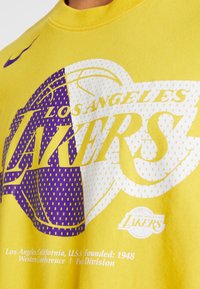 Żółta koszulka z krótkim rękawem z grafiką Los Angeles Lakers. Projekt zawiera fioletowe akcenty, okrągłe logo i tekst o gładkiej fakturze.