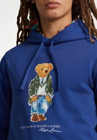 Blå hoodie med en bild av en björn i en grön jacka, vit tröja och jeans med bruna skor. Texten nedanför lyder "POLO BEAR by RALPH LAUREN."