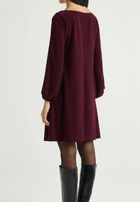 Robe droite bordeaux avec manches longues ballon et encolure ronde. Tissu lisse et coupe évasée, portée avec des bottes noires montantes jusqu'aux genoux.