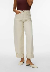 Femme portant un pantalon beige taille haute à jambes larges avec revers, un haut sans manches marron foncé, et des chaussures crème à talons bas et sans lacets.