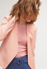 Světle růžový blazer s designem na jedno knoflíkové zapínání, přeložený přes červeno-bílý proužkovaný top. Oblečení doplňují tmavě modré kalhoty s akcentovaným páskem.