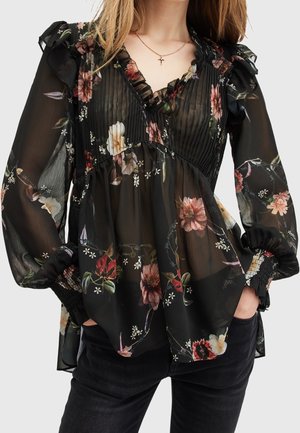 Femme portant un chemisier noir transparent à imprimé floral, poitrine plissée, col et poignets à volants, associé à un pantalon noir et un collier croix.