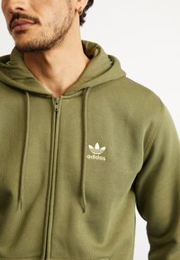 Esguado verde oliva com fecho, feito de material macio, apresentando um fecho frontal, capuz com cordão e logótipo dourado da Adidas no lado esquerdo do peito.