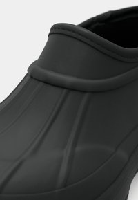 Shoe de borracha preta com superfície texturizada, linhas elevadas na parte superior e um colarinho liso e arredondado. Possui uma sola plana.