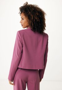 Blazer ajusté et court en tissu texturé magenta avec un dos droit et un détail de couture dans le dos subtil. Assorti avec un pantalon assorti.