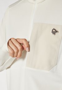 Hoodie v kremni barvi z teksturirano telo, gladko žepom v svetlejši odtenek in logotipom pri žepu. Ima polovično zadrgo.