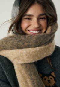 Varm, flerfarvet tørklæde i nuancer af brun og creme, lavet af blødt stof, viklet omkring et smilende ansigt med mellembrunt hår. Mørk sweater nedenunder.