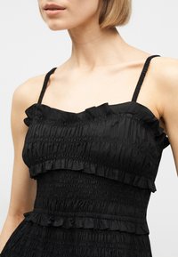 Γυναίκα με μαύρο αμάνικο φόρεμα με ριμπέ ύφασμα και λεπτομέρειες βολάν, που δείχνει τους ώμους και τον λαιμό με φόντο απλό.