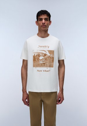 Homme portant un t-shirt blanc avec un élan de dessin animé tenant une carte, texte "Napapijri Lost in Sweden" et "Now what?", associé à un pantalon marron.