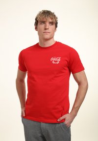 Rød bomuld t-shirt med rund halsudskæring og korte ærmer, med et hvidt Coca-Cola logo trykt på venstre side.