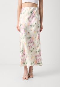 Nainen, jolla on ylhäältä korkea, nilkkapituinen kukallinen satiinihame, jossa on vaaleanpunaisia, vihreitä ja keltaisia kuvioita, sekä beigellä varustetut remmisandalit.