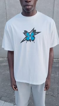 Joven con una camiseta blanca con un gráfico de mariposa azul y negra sobre una estrella negra, de pie frente a un fondo de pared gris.