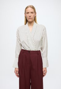 Bijela bluza na preklop s crnim točkicama, dugih rukava. Uparena s visokim strukom vinsko-crvenim hlačama s gumbima i detaljima nabora sprijeda.