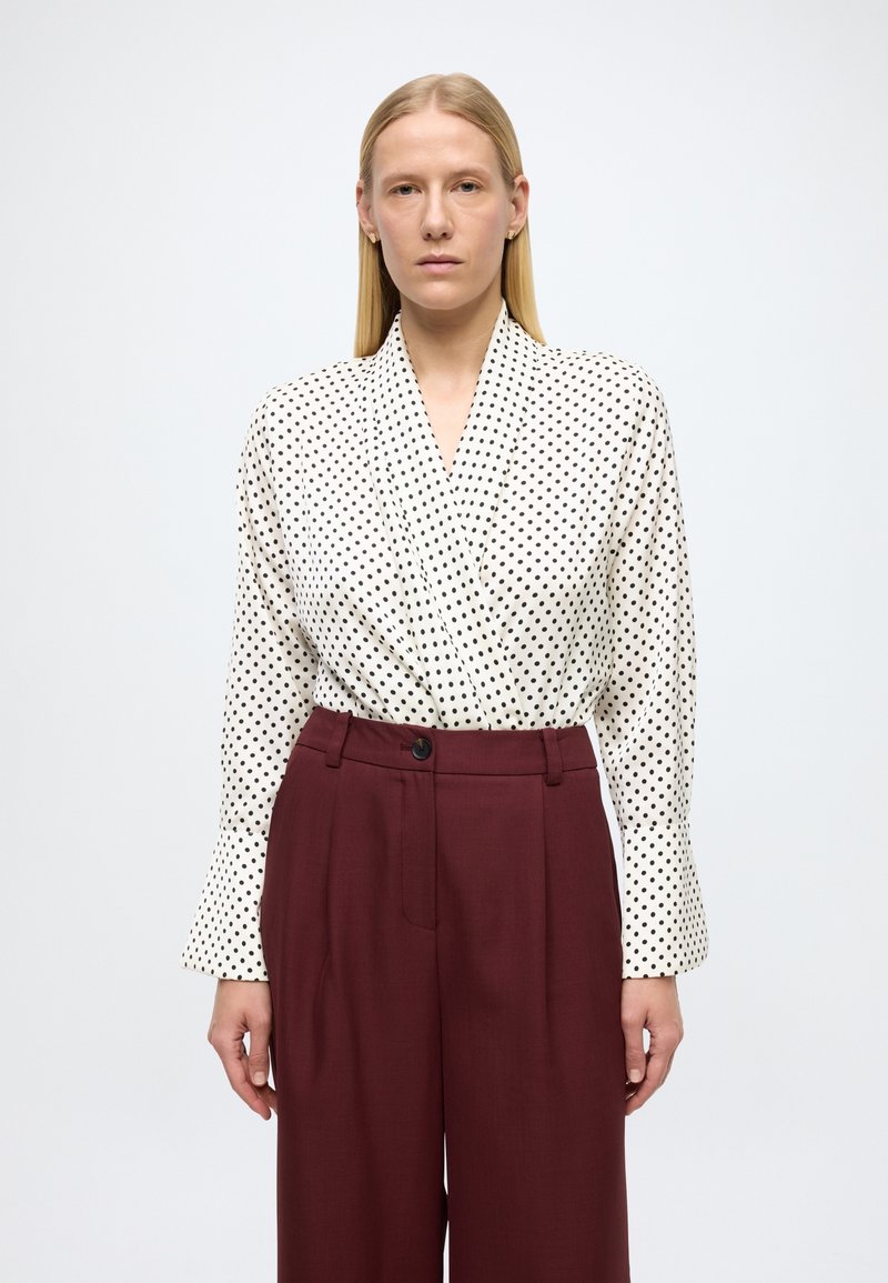 Blusa cruzada blanca con lunares negros, manga larga. Combinada con pantalones burdeos de tiro alto con cierre de botones y detalles de pliegues frontales.