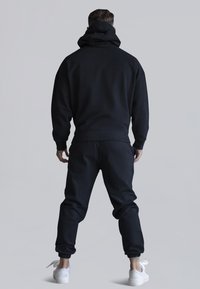 Felpa nera con cappuccio e pantaloni coordinati realizzati in un tessuto morbido e strutturato. Presenta un ampio cappuccio e polsini elastici, abbinati a sneaker bianche.