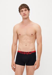 Schwarze Boxershorts mit einem rot-schwarzen elastischen Bund, der das "HUGO"-Logo trägt. Glattes Material, figurbetontes Design und Länge bis zur Mitte des Oberschenkels.