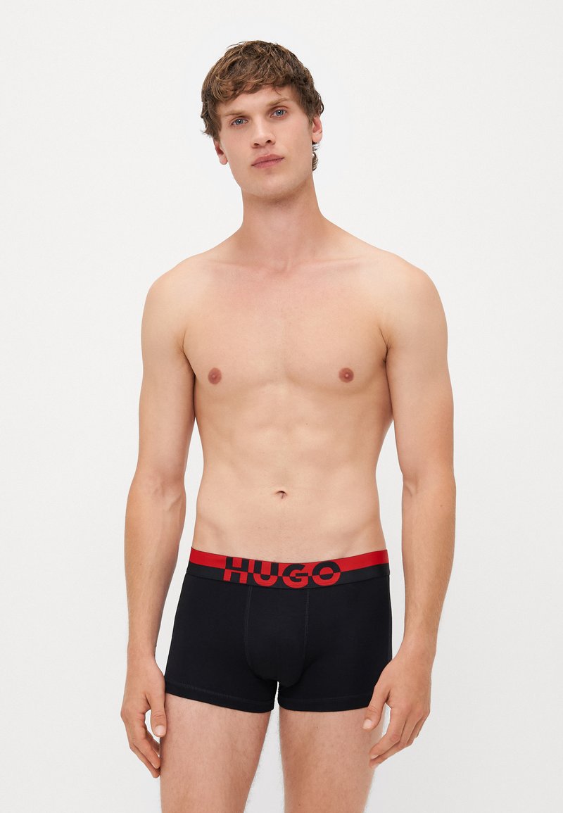 Schwarze Boxershorts mit einem rot-schwarzen elastischen Bund, der das "HUGO"-Logo trägt. Glattes Material, figurbetontes Design und Länge bis zur Mitte des Oberschenkels.