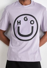 Lavendel bomulds T-shirt med et iøjnefaldende sort cirkulært grafisk design med bogstaverne "HGO" og et smilende ansigt i midten. Korte ærmer.