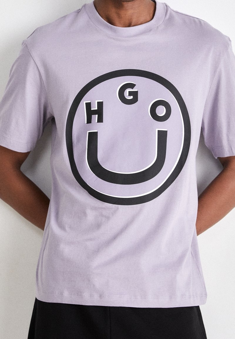Lavendel bomulds T-shirt med et iøjnefaldende sort cirkulært grafisk design med bogstaverne "HGO" og et smilende ansigt i midten. Korte ærmer.