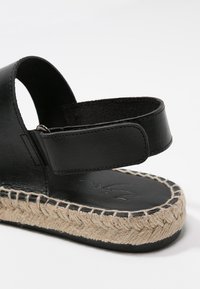 Černá kožená sandál s jute podrážkou ve stylu espadrille, vybavený širokým páskem a nastavitelním suchým zipem, texturovaným povrchem a zaobleným tvarem.