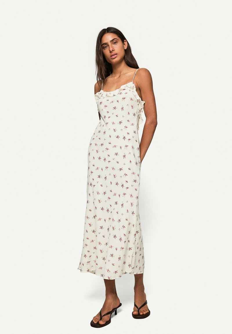 Abito midi floreale color crema con spalline sottili, dettagli a volant lungo il colletto e una silhouette fluida caratterizzata da piccoli fiori rossi.