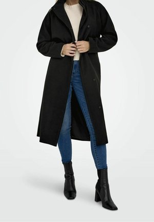 Femme portant un long manteau noir, un pull beige clair, un jean skinny bleu et des bottines noires à talons, debout devant un fond uni.