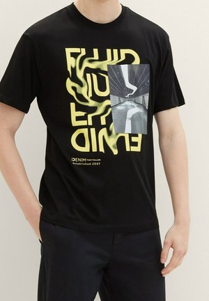 Man wearing zwart T-shirt met gele vervormde letters en een rechthoekige foto van een gebogen witte lijn in een tunnel aan de voorkant.