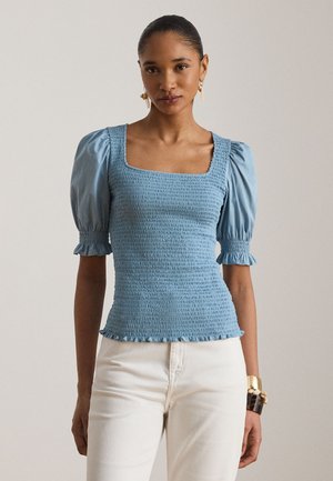 Naine, kellel on kokku tõmmatud juuksed, kannab sobivat sinist krepitud ülaosa puffvarrukate ja valgete pükstega, aksessuaaridena kõrvarõngad ja käevõrud.